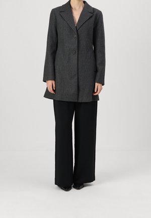Personne portant un manteau gris foncé boutonné avec un col et un pantalon large noir long, se tenant devant un fond clair uni.