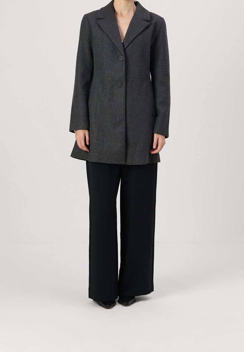 Personne portant un manteau gris foncé boutonné avec un col et un pantalon large noir long, se tenant devant un fond clair uni.
