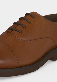 Chaussure Oxford en cuir marron avec des lacets marron foncé et un talon bas, montrant des coutures détaillées et une finition lisse.