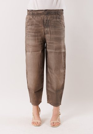 Bruine losse broek met elastische tailleband, gedragen met een wit topje en beige sandalen met open teen en hak, op een lichte achtergrond.