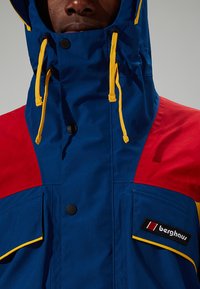 Bunte Jacke mit einem blauen Körper, roten Schultern, gelben Akzenten, einem hohen Kragen, Kordelzügen und einem Logopatch am linken Arm.