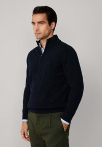 Maglione blu navy a maglia cable con colletto alto, dotato di una zip a mezza lunghezza. Indossato sopra una camicia a colletto azzurro e pantaloni verdi.