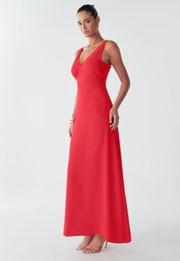 Täismõõdus kleit sügav punane, sügava V-kaelusega ja keha järgiva ülakehaga. Valmistatud siledast kangast, millel on voolav, kuni pahkluud ulatuv seelik.