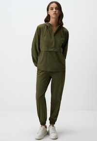 Survêtement vert olive avec un haut à capuche zippé à demi et un pantalon fuselé. Il comprend des poignets élastiques et des poches latérales zippées, fabriqué en tissu léger.