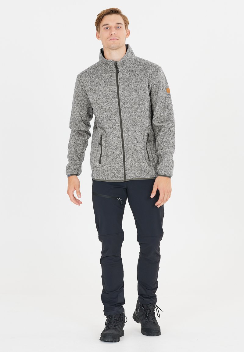 Grå fleecejakke med høj krave, lynlås og to sidelommer, parret med sorte bukser og vandrestøvler. Glat tekstur, holdbart design.