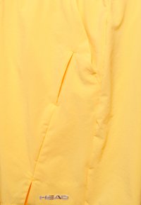 Gelbe Sportjacke mit glatter Textur, ausgestattet mit schrägen seitlichen Taschen und einem silbernen "HEAD"-Logo unten rechts.