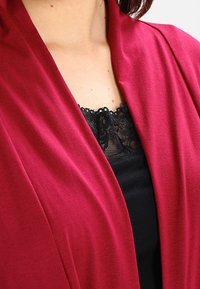 Bordový zavinovací kardigan s měkkou, hladkou texturou a splývavým designem, vrstvený přes černou tílko s krajkovým lemem.