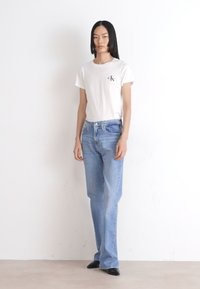 Camiseta blanca de algodón con un pequeño logo negro, combinada con unos vaqueros acampanados de color azul claro. El modelo lleva zapatos negros de punta en un fondo sencillo.