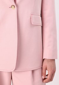 Blazer rose clair ajusté avec un bouton unique, une poche à rabat et des surpiqûres contrastantes sur les bords. Tissu texturé.