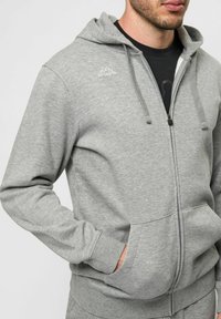 Felpa grigia con zip realizzata in tessuto morbido, dotata di tasca a marsupio, cappuccio con cordini e logo ricamato sul lato sinistro del petto.