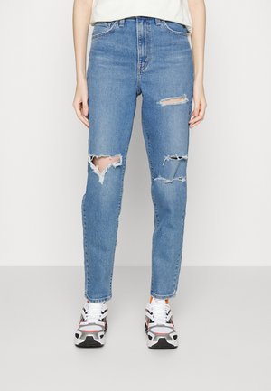Personne portant un jean bleu clair déchiré et des baskets blanches, noires et orange, debout devant un fond clair uni.