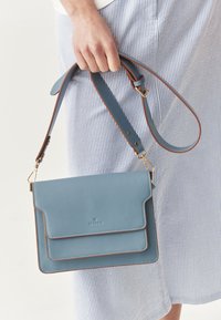 Svijetloplava kožna torbica na rame s dva prednja džepa, smeđim naglascima, podesivim remenom i suptilnim brendiranjem sprijeda.