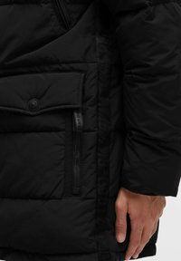 Schwarze Pufferjacke aus strukturiertem Stoff, mit einer großen Klapptasche mit Knopf und einer seitlichen Reißverschlusstasche, die ein elegantes Design betont.