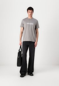 Camiseta de algodón gris con logo blanco de "Calvin Klein", combinada con jeans de pierna ancha negros, zapatos negros y un bolso tote negro.