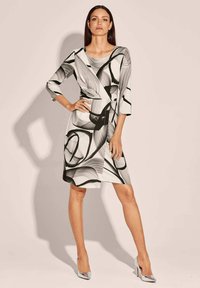 Schwarz-weißes Kleid mit wirbelndem abstraktem Muster, dreiviertellange Ärmel, Knotendetail an der Taille, knielang, kombiniert mit metallischen High Heels.