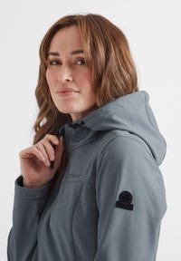 Grey hooded jacket made of soft, textured material featuring a sleek design, logo on the sleeve, and cinch cords for adjustable fit.