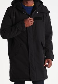 Mann trägt einen langen schwarzen Wintermantel mit Kapuze und schließt den Reißverschluss vorne über einem blauen Pullover mit gelbem Kragen.