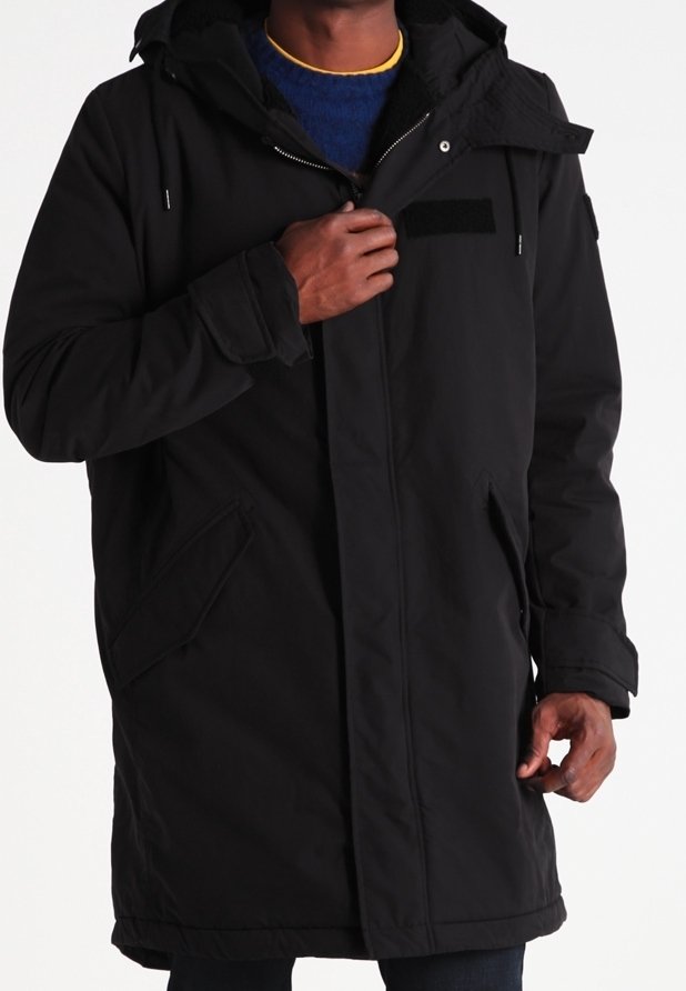 Mann trägt einen langen schwarzen Wintermantel mit Kapuze und schließt den Reißverschluss vorne über einem blauen Pullover mit gelbem Kragen.