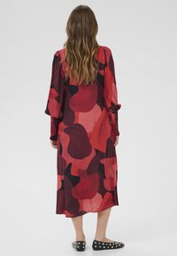 Langes, fließendes Kleid in Rot und Burgunder mit einem abstrakten Muster. Leichter Stoff, lange Ärmel und lockerer Schnitt. Akzentuiert durch schwarze flache Schuhe mit Nieten.