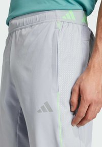 Jasnoszare szorty sportowe o teksturowanym wzorze, z zielonymi akcentami, elastycznym paskiem w talii i logo Adidas po stronie.