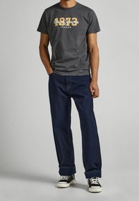 Hombre vistiendo una camiseta oscura gris de Pepe Jeans London 1973, pantalones vaqueros oscuros de mezclilla doblados en los puños y zapatillas negras y blancas, de pie con las manos en los bolsillos.