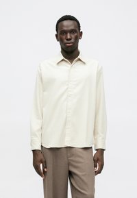Chemise à manches longues de couleur crème avec un devant à boutons, présentant une texture côtelée et un col classique rabattu, associée à un pantalon marron clair.