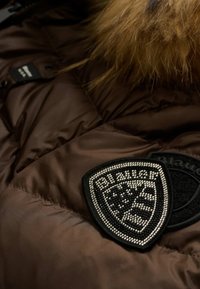 Tessuto marrone imbottito per giacca con una texture liscia e lucida. Presenta una toppa nera ricamata con dettagli argentati e un bordo collo in pelliccia.