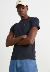 Tricou negru din bumbac cu mânecă scurtă, având un buzunar pe piept și detalii cu logo. Purtați cu blugi decolorati de culoare albastră deschisă și o șapcă bej.