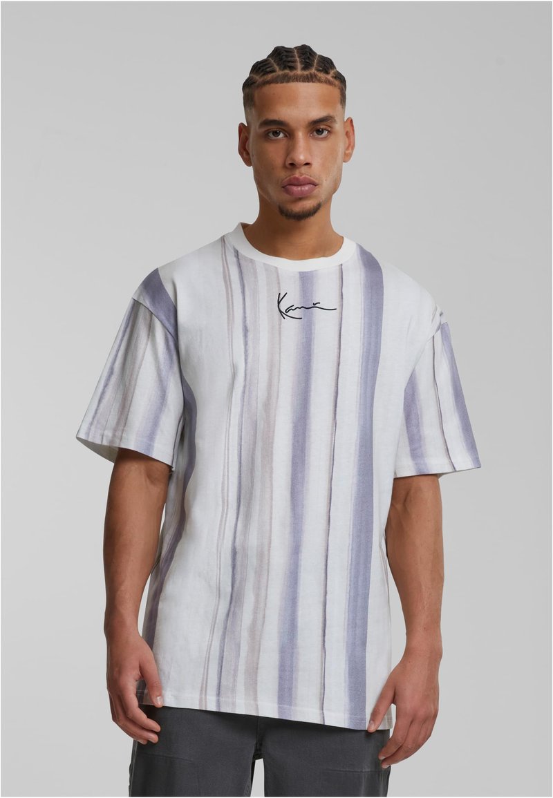 Férfi, akinek fonott haja van, fehér és szürke függőleges csíkos pólót visel, amelynek mellkasán egy kis fekete aláírásos logó található.