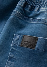 Blå denimbyxor med elastisk midjeband, med en svart läderetikett på bakfickan. Texturen visar ett blekt tvättmönster.