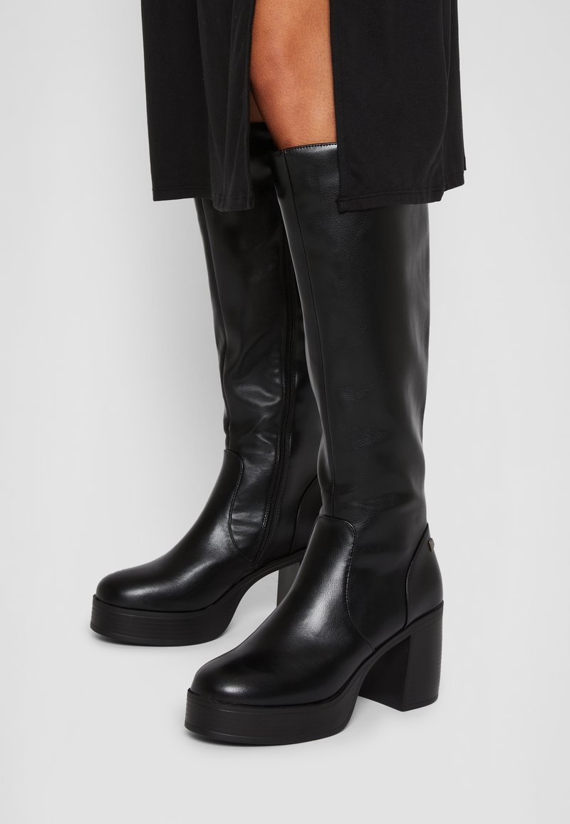 Bottes en cuir noires montantes jusqu'au genou avec talons carrés et semelles plateformes, portées avec une robe noire.
