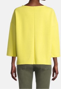 Geel oversized sweatshirt met een brede halslijn en driekwart mouwen, gemaakt van zachte stof. Getextureerde stiksels langs de achterkant.