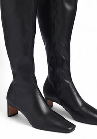 Čierne kožené čižmy po kolená s špicatou špičkou, so bočným zipsom a blokovým podpätkom s hnedým akcentom. Hladká textura.