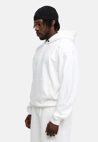 White cotton hoodie with a front pocket, adjustable drawstring hood, and ribbed cuffs. Paired with matching sweatpants, worn by a person.