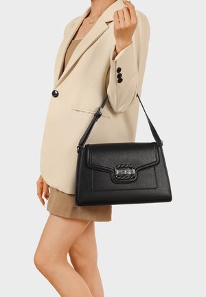 Donna con blazer beige e gonna che tiene una borsa a tracolla in pelle nera con fibbia d'argento su una spalla, su sfondo bianco.