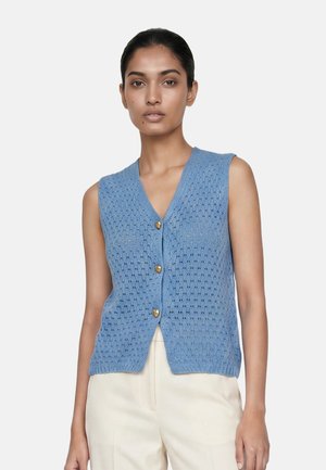 Donna che indossa un gilet di maglia blu senza maniche con tre bottoni dorati a forma di cuore e pantaloni color avorio, in piedi davanti a uno sfondo semplice.