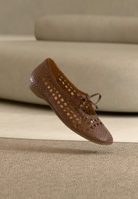 Chaussure plate en cuir tressé marron avec des détails découpés et un nœud sur le dessus, flottant au-dessus d'un tapis texturé avec un canapé beige en arrière-plan.