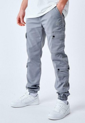 Pantalones cargo grises hechos de una tela suave, con bolsillos laterales, puños elásticos y un logo bordado en el muslo. Llevados con zapatillas blancas.