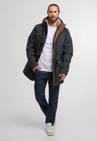 Parka bleu foncé avec des accents marron, une capuche doublée en fausse fourrure, plusieurs poches et des fermetures éclair. Porté sur une chemise blanche et un pantalon marine.