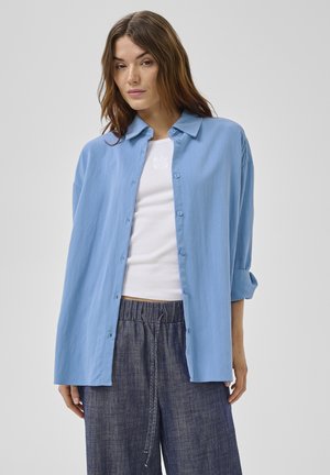 Jonge vrouw die een loszittend lichtblauw overhemd draagt over een witte tanktop en donkergrijze broek met aantrekkoord, staand tegen een grijze achtergrond.