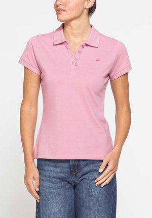 Donna che indossa una polo rosa aderente a maniche corte con bottoni e jeans blu, in piedi davanti a uno sfondo bianco.