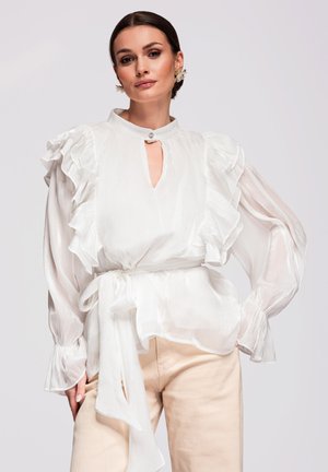 Kobieta w białej bluzce z falbankami na długich rękawach i dekoltem w kształcie łezki oraz beżowych spodniach, pozująca na tle jasnego, jednolitego tła.