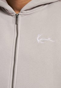 Lys grå hettejakke med en jevn tekstur, med en hvit brodert logo på brystet og et standard hette-design.