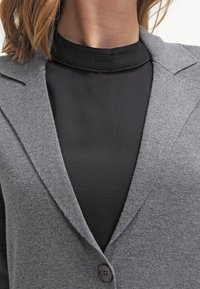 Šedý kardigan s texturovaným povrchem, se šálovým límcem a jedním černým knoflíkem, vrstvený přes černý top s vysokým výstřihem.