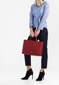 Sac fourre-tout en cuir rouge avec deux poignées, texture lisse et un logo sur le devant. Associé à une chemise rayée et un pantalon à rayures fines.