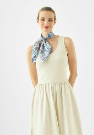 Femme aux cheveux clairs, vêtue d'une robe crème sans manches et portant un foulard en soie bleu à motifs noué autour du cou.