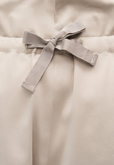 Detailný záber béžových látkových nohavíc s viazanou mašličkou z grosgrainovej stuhy na páse a viditeľnými detailmi šitia.