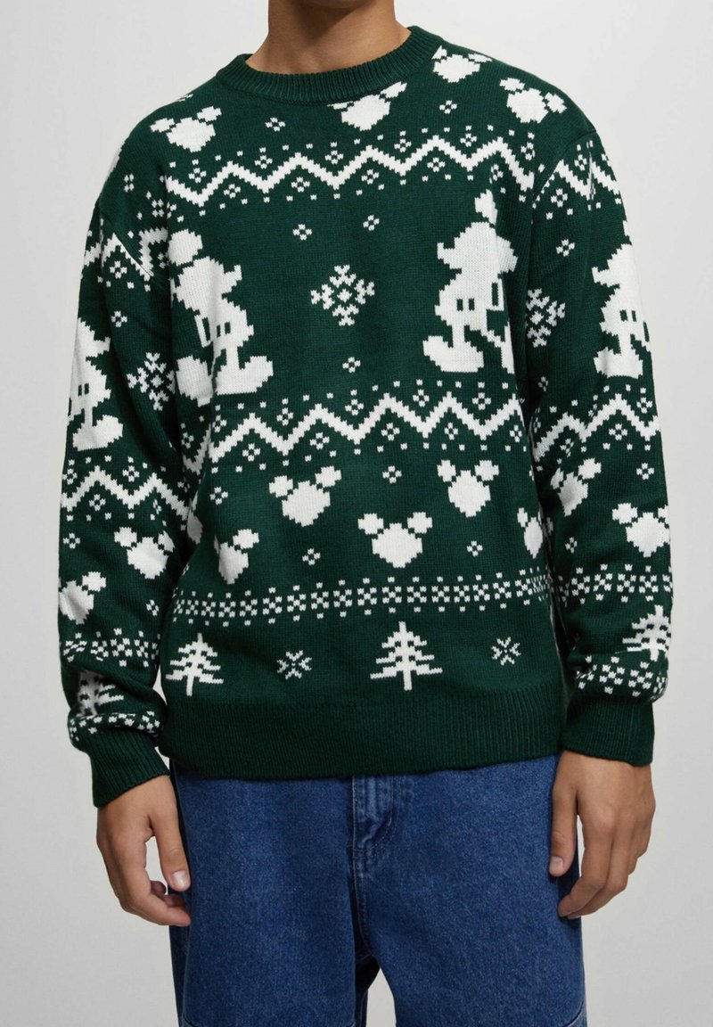 Pull vert avec des figures de dessins animés pixelisées blanches, des cœurs, des flocons de neige et des motifs de sapins de Noël porté avec un jean bleu.