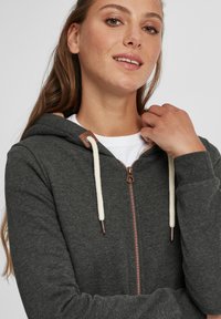 Sweat à capuche zippé gris avec une capuche en polaire, présentant des cordons crème et une fermeture éclair en bronze. Tissu doux et design ajusté.