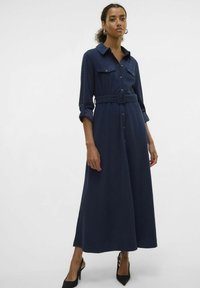 Robe bleu marine à manches longues, ceinture à la taille, deux poches poitrine et jupe évasée. Portée avec des talons noirs pointus.